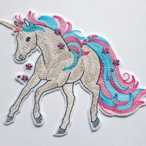 Könnte beinhalten: Gestickter Aufnäher mit einem weißen Einhorn mit blauem und rosafarbenem Mähne und Schweif. Das Einhorn hat ein goldenes Horn und ist mit rosa Blumen verziert.