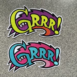 Könnte beinhalten: Zwei gestickte Aufnäher mit dem Wort "GRRR!" in einer Comic-Schriftart. Der obere Aufnäher ist gelb, lila und orange. Der untere Aufnäher ist blau, rosa und türkis.