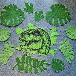 Könnte beinhalten: Grüne Filzpatches in Form von Blättern und Dinosaurierspuren, mit einer Schwarzweißillustration eines Dinosaurierkopfes in der Mitte.