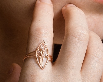 Könnte beinhalten: Ein zarter Goldring mit einem Blattdesign. Der Ring wird am Finger einer Person mit roten Haaren getragen.