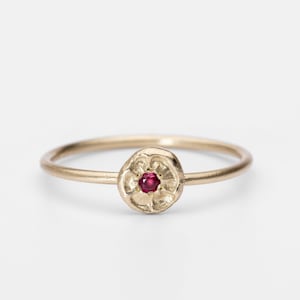以下が含まれることがあります： 繊細なゴールドリング、花のモチーフ。リングの中央には、小さな丸い赤い宝石があります。バンドは細く滑らかで、リングは白い背景にセットされています。