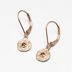 Op de afbeelding: Gouden oorbellen met een kleine roze edelsteen in een ronde hanger met een bloemmotief.