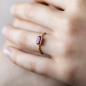 Pode incluir: Um anel de ouro com uma pedra vermelha retangular. O anel está no dedo de uma pessoa.