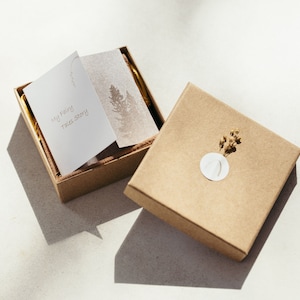 K&ouml;nnte beinhalten: Eine braune Pappbox mit einer wei&szlig;en Karte im Inneren, auf der "My Fairy Tale Story" steht. Die Box ist ge&ouml;ffnet und enth&auml;lt eine kleinere braune Box. Die kleinere Box hat einen wei&szlig;en Kreis mit einem Design und einen kleinen Zweig getrockneter Blumen.