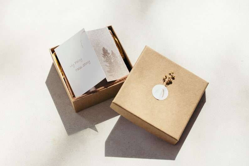 K&ouml;nnte beinhalten: Eine braune Pappbox mit einer wei&szlig;en Karte im Inneren, auf der "My Fairy Tale Story" steht. Die Box ist ge&ouml;ffnet und enth&auml;lt eine kleinere braune Box. Die kleinere Box hat einen wei&szlig;en Kreis mit einem Design und einen kleinen Zweig getrockneter Blumen.