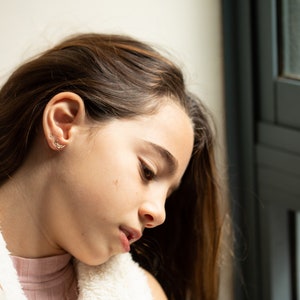 K&ouml;nnte beinhalten: Eine junge Person mit langen braunen Haaren schaut nach unten. Sie tr&auml;gt eine wei&szlig;e flauschige Jacke und ein rosa Hemd. Die Person steht in der N&auml;he eines Fensters.