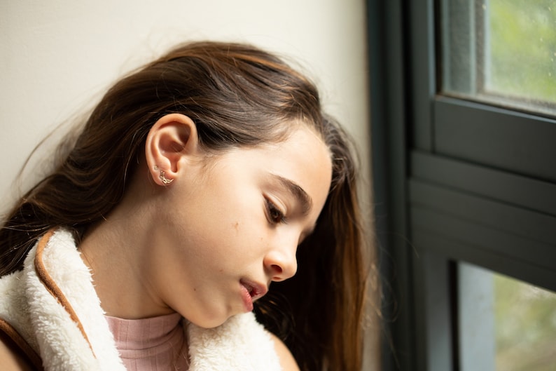 K&ouml;nnte beinhalten: Eine junge Person mit langen braunen Haaren schaut nach unten. Sie tr&auml;gt eine wei&szlig;e flauschige Jacke und ein rosa Hemd. Die Person steht in der N&auml;he eines Fensters.