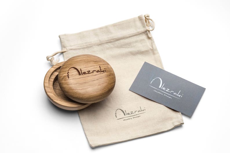 May include: A wooden jewelry box with a lid, open to reveal the interior. The box is next to a drawstring bag and a gray business card. The text "Alezraki" is engraved on the box and printed on the bag and card.