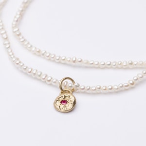 May include: A delicate white pearl necklace with a gold charm featuring a red gemstone in the center. The charm is shaped like a flower.
