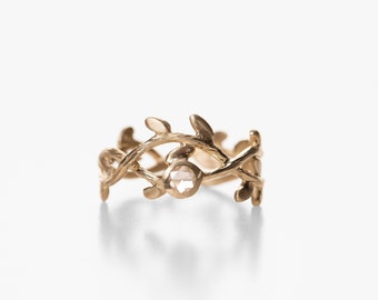 May include: A gold ring featuring a delicate, intricate design of intertwined branches and leaves. A clear, faceted gemstone is set in the centre of the ring.
