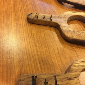 Może przedstawiać: Trzy drewniane podkładki pod napoje z uchwytem. Każda podkładka ma wygrawerowane na uchwycie inicjały "K + L".