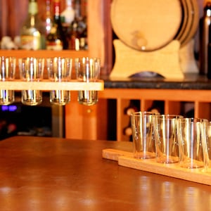 May include: A wooden serving tray with six clear glass shot glasses. The tray is being held by a hand and the glasses are filled with a clear liquid. The tray is being placed on a wooden table.
