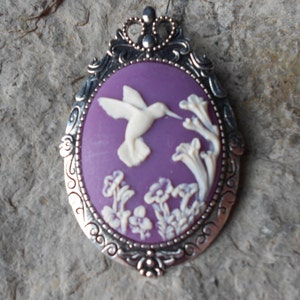 May include: A silver cameo pendant with a purple background and a white cameo of a hummingbird in flight. The hummingbird is surrounded by white flowers.