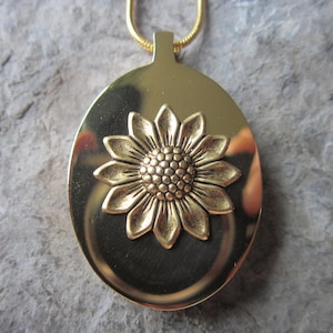 May include: Gold oval pendant necklace featuring a sunflower design. The sunflower is raised and has a textured centre.