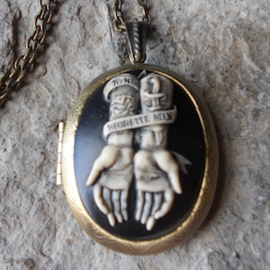 May include: A gold oval locket necklace with a black enamel background and a white cameo of two hands clasped together. The hands are holding a banner with the text "TO ME  HE  HIGHEST  HIX".