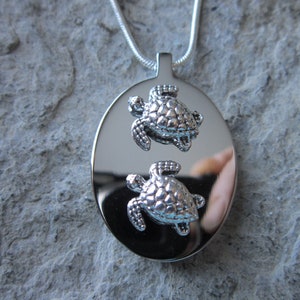May include: Silver pendant necklace featuring two silver turtle charms. The pendant is oval shaped and has a polished finish.
