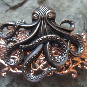 Könnte beinhalten: Eine schwarze Kraken-Haarklammer mit filigranen Details in Roségold. Der Krake hat acht Tentakel mit Saugnäpfen und großen Augen. Die Klammer ist aus Metall und dazu bestimmt, das Haar zu fixieren.
