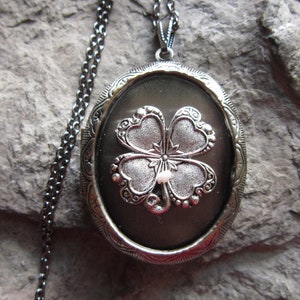 Peut inclure: Un collier avec un médaillon en argent en forme de trèfle à quatre feuilles. Le médaillon est de forme ovale et a un fond noir. Le trèfle est en argent et a une petite perle blanche au centre.