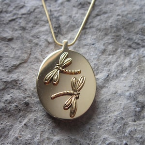 May include: A gold necklace with a pendant featuring two dragonfly designs. The pendant is oval shaped and the dragonflies are embossed on the front.