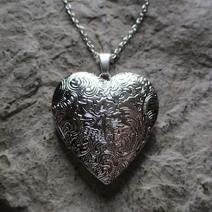May include: Silver heart-shaped locket necklace with intricate floral and swirl pattern. The pendant hangs from a delicate chain. The locket is displayed against a textured, gray stone background.