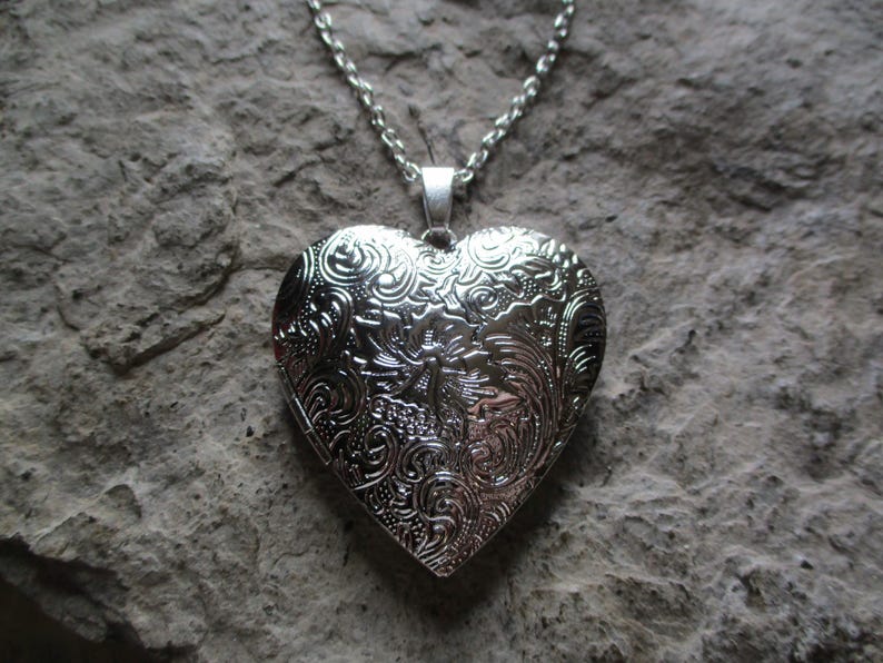 May include: Silver heart-shaped locket necklace with intricate floral and swirl pattern. The pendant hangs from a delicate chain. The locket is displayed against a textured, gray stone background.