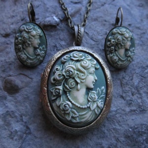 Pode incluir: Um conjunto de colar e brincos com um camafeu de estilo vintage. O pingente apresenta um camafeu branco de um perfil de mulher sobre um fundo verde. Os brincos s&atilde;o semelhantes, com camafeus menores. As joias s&atilde;o feitas de metal com acabamento bronze.