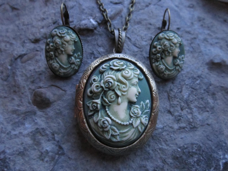 Pode incluir: Um conjunto de colar e brincos com um camafeu de estilo vintage. O pingente apresenta um camafeu branco de um perfil de mulher sobre um fundo verde. Os brincos s&atilde;o semelhantes, com camafeus menores. As joias s&atilde;o feitas de metal com acabamento bronze.