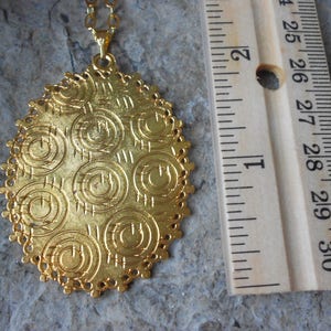 May include: Gold-toned pendant necklace with an oval, openwork design. The pendant features a textured surface with repeating circular patterns. The necklace is displayed with a wooden ruler for scale, showing the pendant's approximate size.