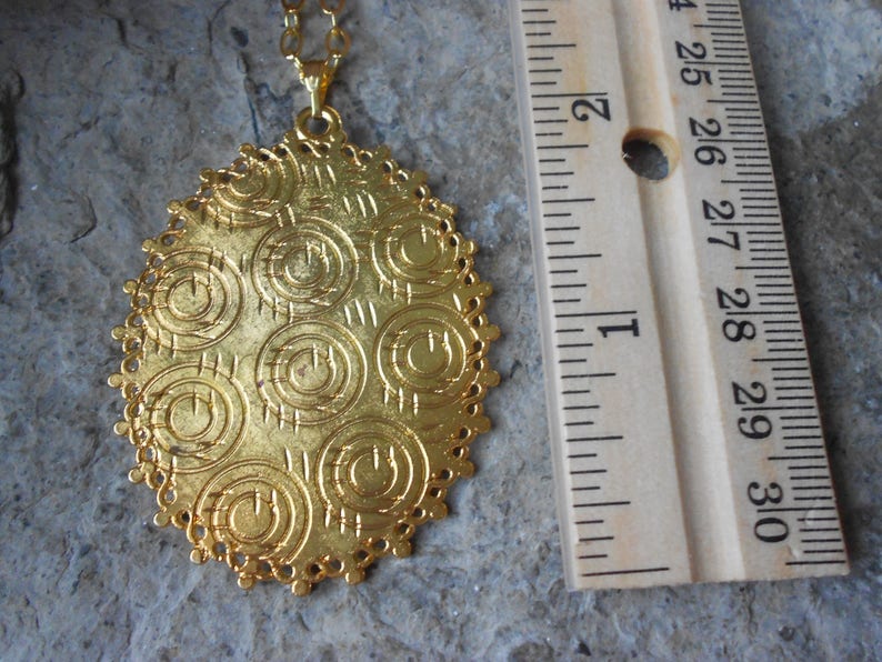 May include: Gold-toned pendant necklace with an oval, openwork design. The pendant features a textured surface with repeating circular patterns. The necklace is displayed with a wooden ruler for scale, showing the pendant's approximate size.
