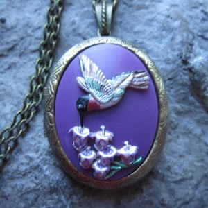 Peut inclure: Un collier avec un médaillon violet orné d'un colibri détaillé. Le colibri est représenté avec son bec dans un groupe de fleurs violettes. Le médaillon est sur une chaîne en bronze.