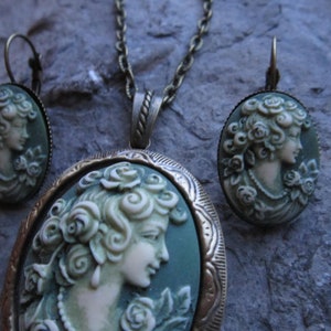 Pode incluir: Um conjunto de colar e brincos de camafeu verde com um retrato detalhado de uma mulher com rosas no cabelo. As joias s&atilde;o de bronze e t&ecirc;m um estilo vintage.