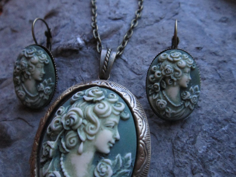 Pode incluir: Um conjunto de colar e brincos de camafeu verde com um retrato detalhado de uma mulher com rosas no cabelo. As joias s&atilde;o de bronze e t&ecirc;m um estilo vintage.