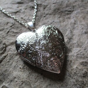 May include: A silver heart-shaped locket necklace with an ornate floral design. The pendant is suspended from a delicate chain. The locket is closed, and the surface is reflective, set against a textured gray background.