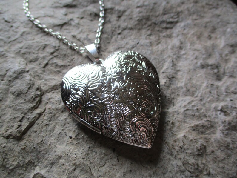 May include: A silver heart-shaped locket necklace with an ornate floral design. The pendant is suspended from a delicate chain. The locket is closed, and the surface is reflective, set against a textured gray background.