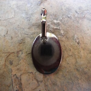 Puede incluir: Pendiente de metal plateado con forma redonda y plana. El pendiente tiene un bucle en la parte superior para sujetarlo a una cadena o cord&oacute;n.