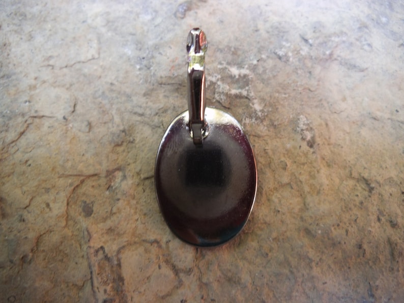 Puede incluir: Pendiente de metal plateado con forma redonda y plana. El pendiente tiene un bucle en la parte superior para sujetarlo a una cadena o cord&oacute;n.