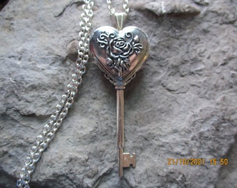 May include: Silver-toned key pendant necklace. The pendant is heart-shaped with a rose design and a key shape. The necklace chain is also silver-toned. The key pendant is displayed on a textured, gray surface.
