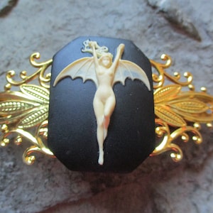 Puede incluir: Una pinza para el pelo dorada con un camafeo negro que presenta una figura esculpida en blanco de una mujer con alas. El camafeo está rodeado por un diseño de filigrana dorada.