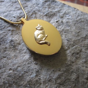 Op de afbeelding: Gouden ovale ketting met een gedetailleerd kattenontwerp. De kat zit met zijn staart om zijn lichaam gekruld.