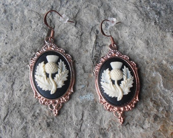 May include: A pair of rose gold earrings with black and white cameo designs featuring a stylized thistle flower.