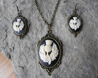 May include: A set of jewelry featuring a black and white cameo pendant and matching earrings. The cameo features a white thistle flower design on a black background. The pendant and earrings are set in an ornate bronze frame.