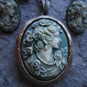 Pode incluir: Um colar com um medalh&atilde;o de camafeu verde adornado com um camafeu branco que representa uma mulher com rosas no cabelo. O medalh&atilde;o &eacute; rodeado por uma moldura de metal dourado.