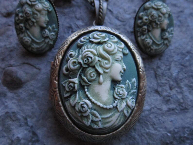 Pode incluir: Um colar com um medalh&atilde;o de camafeu verde adornado com um camafeu branco que representa uma mulher com rosas no cabelo. O medalh&atilde;o &eacute; rodeado por uma moldura de metal dourado.