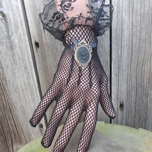 May include: Black lace gloves with a black and gold cameo brooch. The gloves are made of a fine mesh fabric and have a delicate floral pattern.
