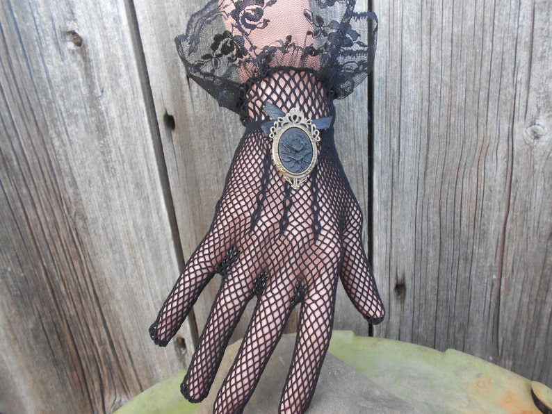 May include: Black lace gloves with a black and gold cameo brooch. The gloves are made of a fine mesh fabric and have a delicate floral pattern.