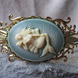 May include: A gold-tone bracelet with a blue and white cameo featuring a mermaid. The cameo is surrounded by a decorative filigree design.