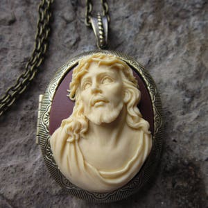 Può includere: Una collana con un ciondolo dorato con un cammeo di un volto maschile. Il cammeo è realizzato in porcellana bianca e raffigura un uomo con i capelli lunghi e la barba. Il ciondolo è di forma ovale e presenta un design dettagliato attorno al bordo.