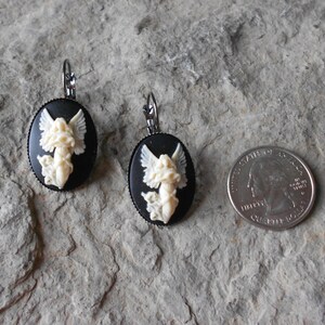 Puede incluir: Un par de pendientes de camafeo negro y blanco con un dise&ntilde;o de &aacute;ngel blanco. Los pendientes son de forma ovalada y tienen un cierre de gancho plateado.