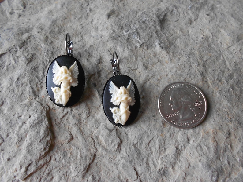 Puede incluir: Un par de pendientes de camafeo negro y blanco con un dise&ntilde;o de &aacute;ngel blanco. Los pendientes son de forma ovalada y tienen un cierre de gancho plateado.