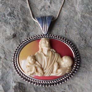Puede incluir: Collar con colgante ovalado con un relieve crema de una figura con dos niños, sobre un fondo rojo y amarillo. El colgante tiene un borde con cuentas plateadas y cuelga de una cadena plateada.